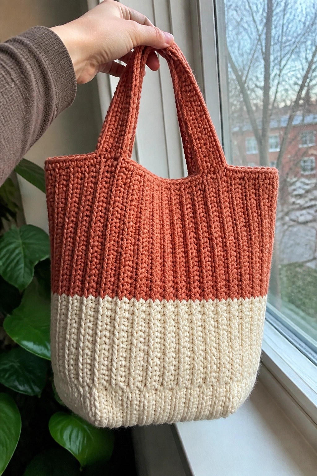 Hand holding orange and cream ribbed knit tote bag by window with plants