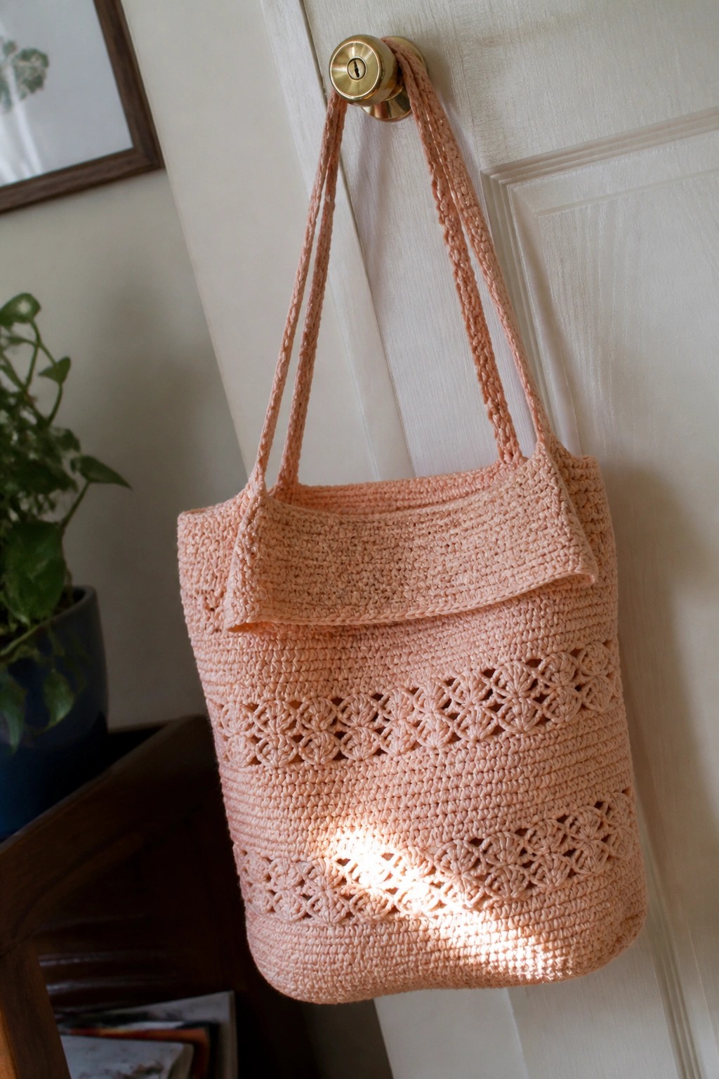 Peach crocheted tote bag with lace pattern hanging from brass doorknob on white door