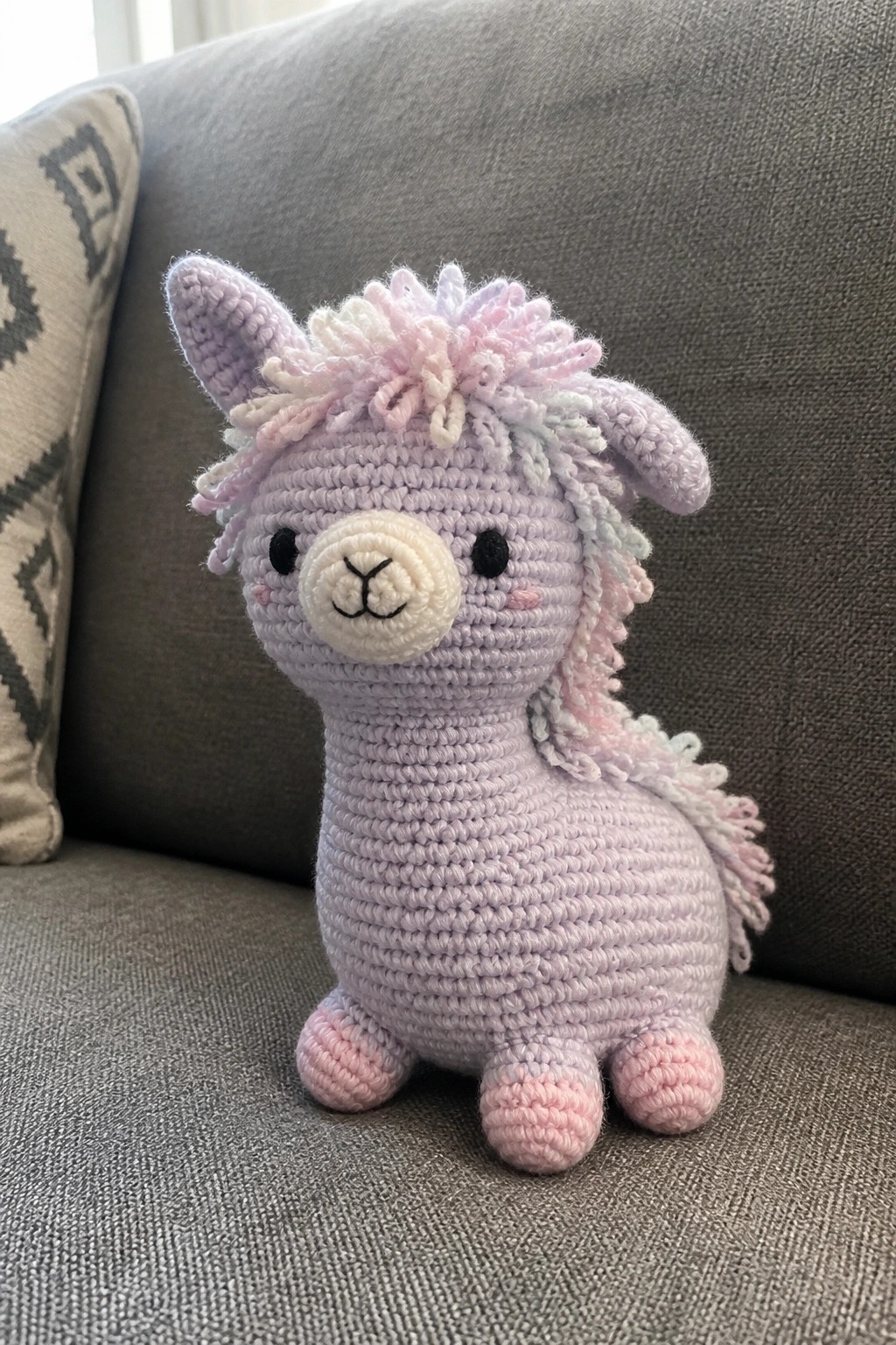 Crocheted lavender llama toy with pink-and-white mane sits on gray couch.
