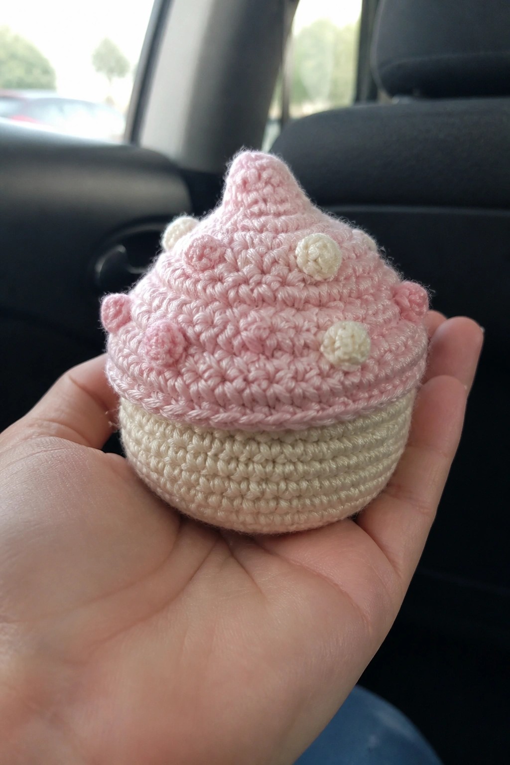 Hand holding pink crocheted cupcake with white dots and beige base in car