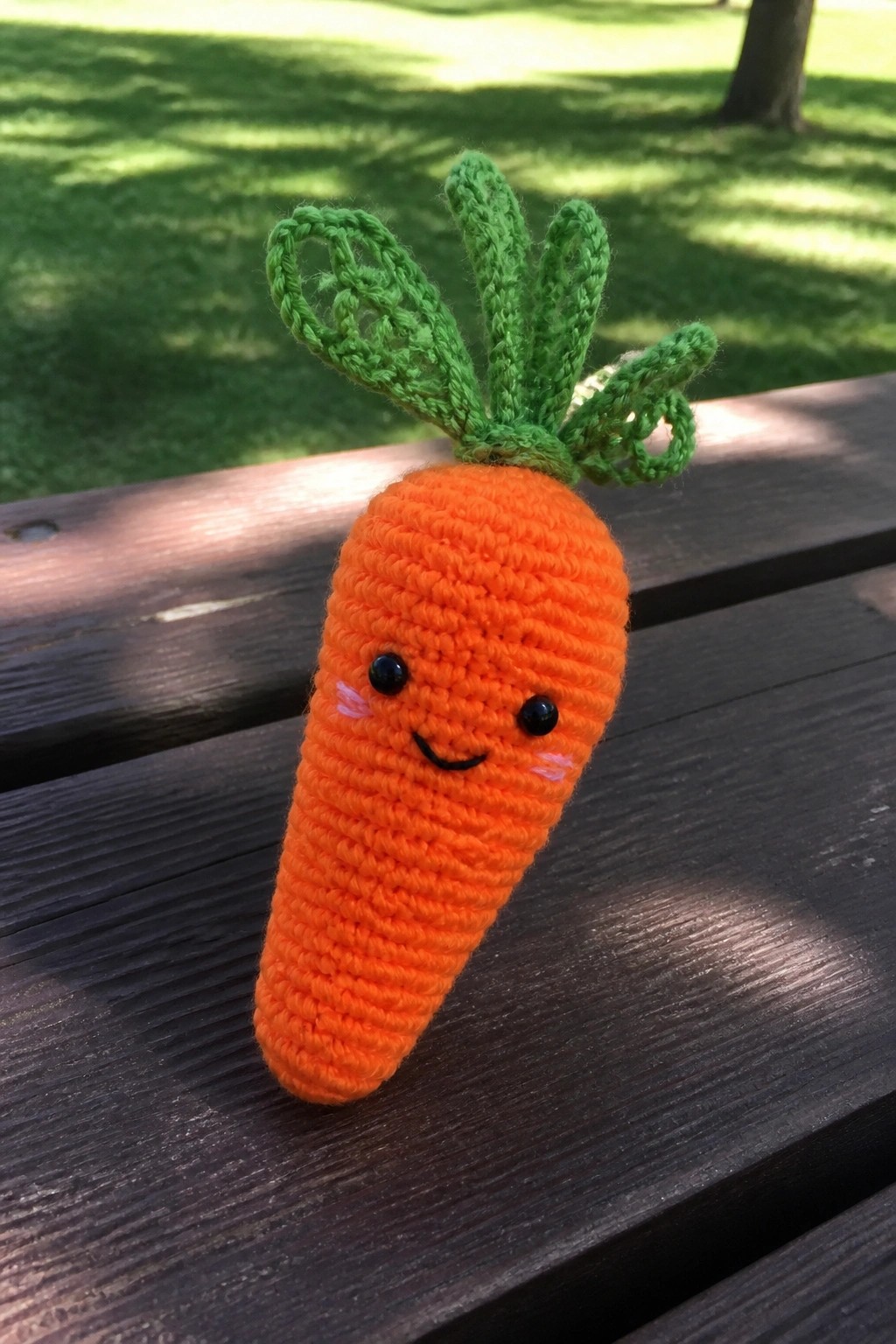 Crocheted orange carrot with smiling face and green leaves on wooden bench outdoors
