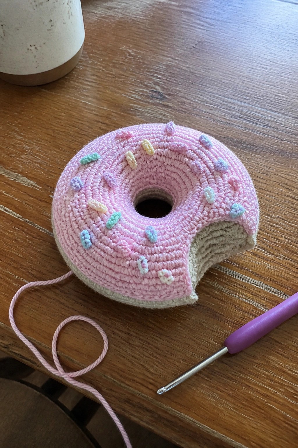 Pink crocheted donut with pastel sprinkles and missing bite beside purple crochet hook on wood table.