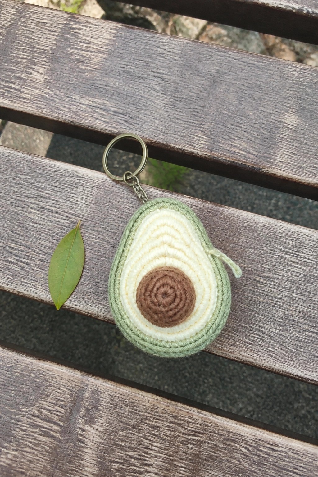 Crocheted avocado keychain on wooden bench beside small green leaf