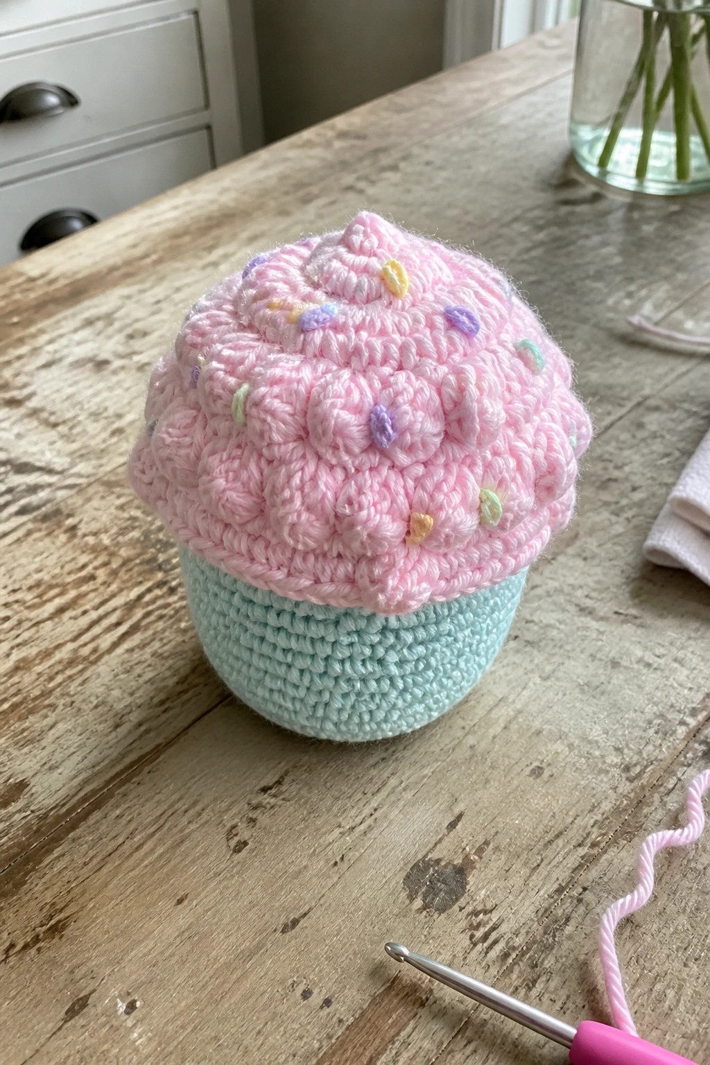A pink crocheted cupcake with multicolored dots sits on a wooden table beside a crochet hook.
