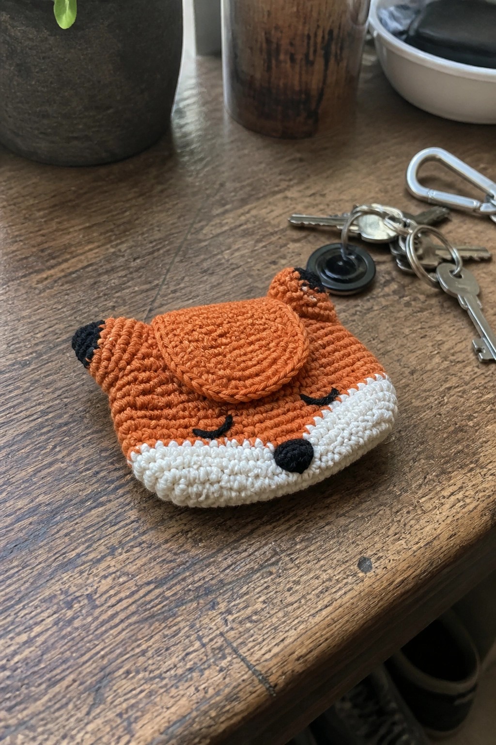 A small orange crocheted fox pouch with white muzzle, black nose and ear tips, and closed eyes rests on a wooden table beside a set of keys.