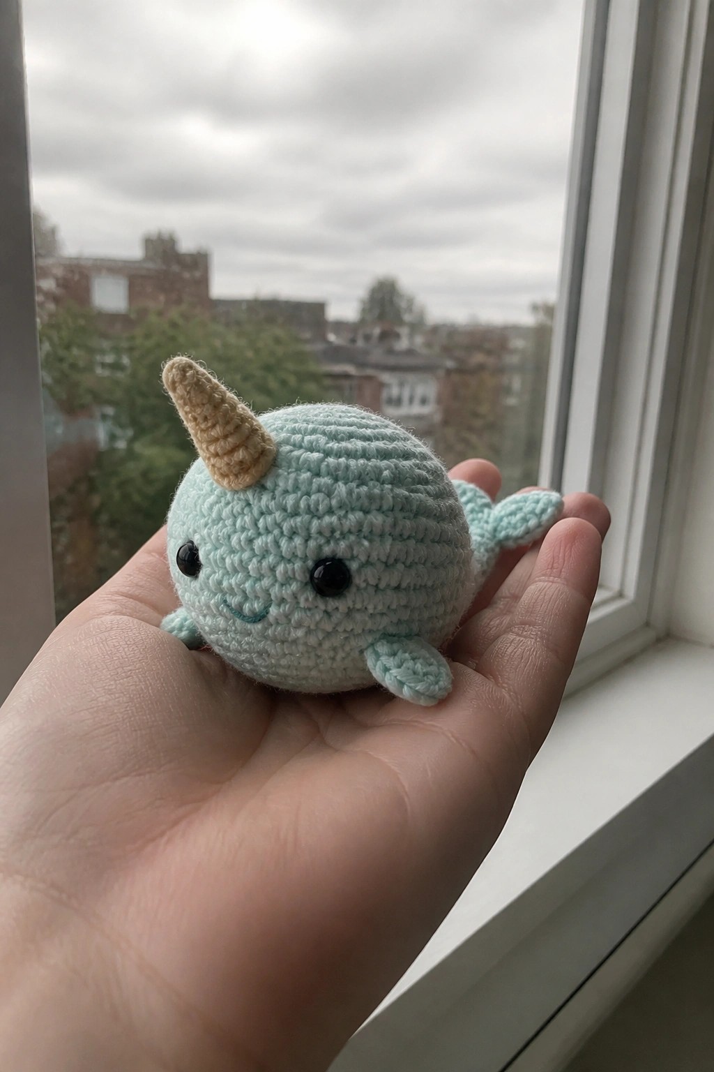 Hand holding light blue crocheted narwhal with beige horn near window.