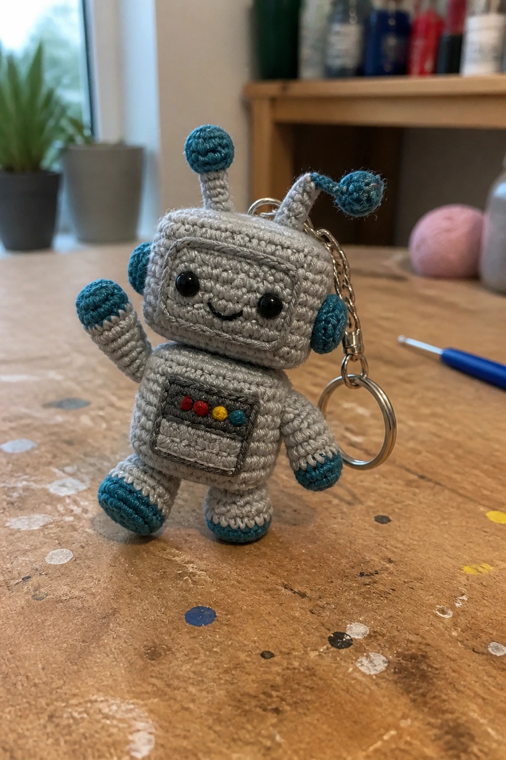 Crocheted gray robot keychain with teal accents and raised arm on wood table
