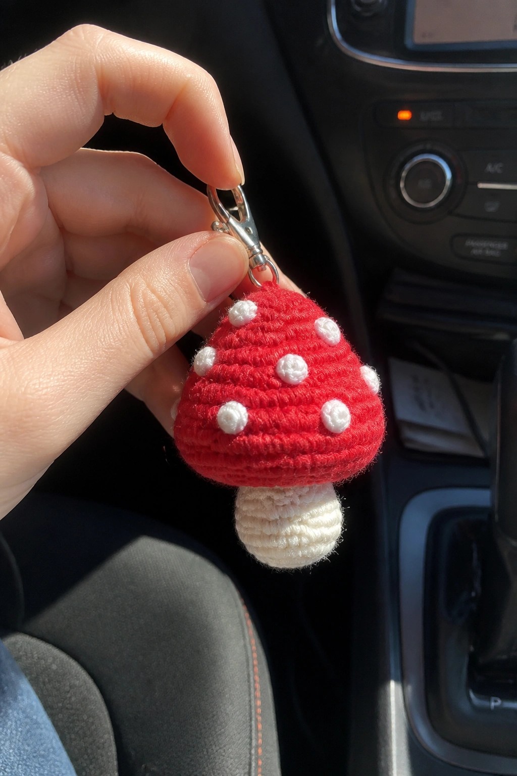 Hand holding crocheted red mushroom keychain with white spots in car interior
