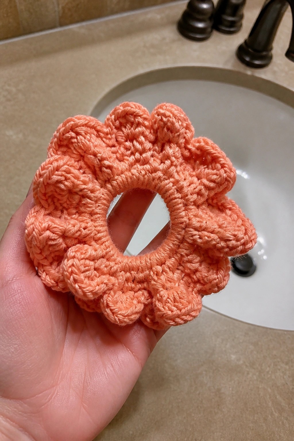 Hand holding a peach crocheted ruffled scrunchie in front of a bathroom sink