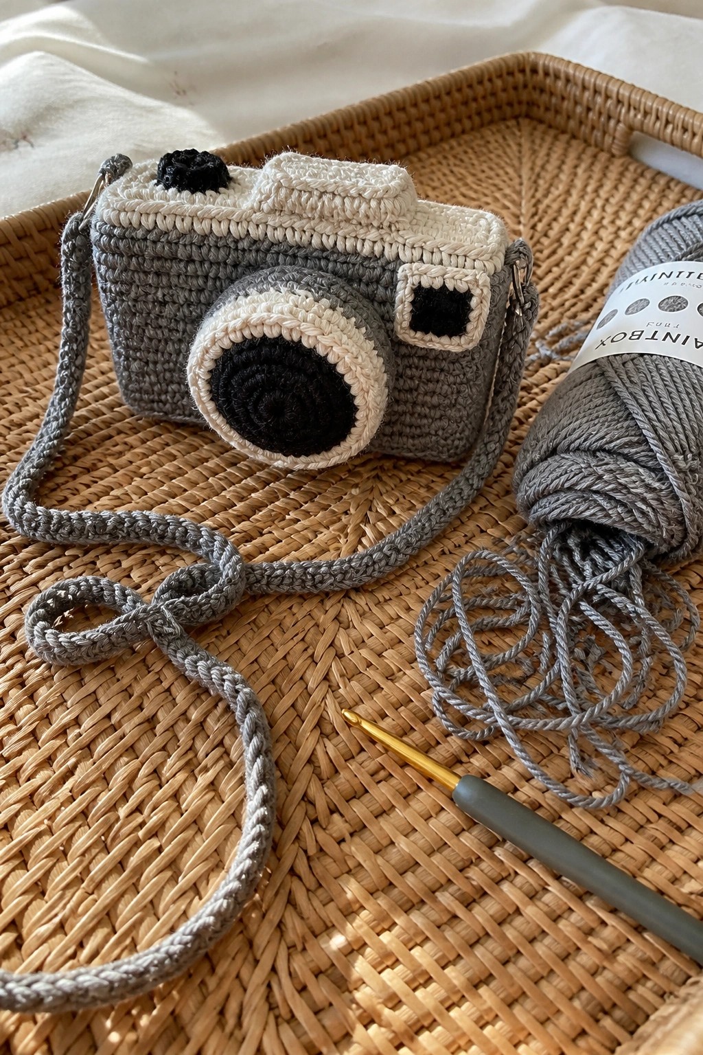 Crocheted gray camera bag with yarn skein and hook on woven rattan tray