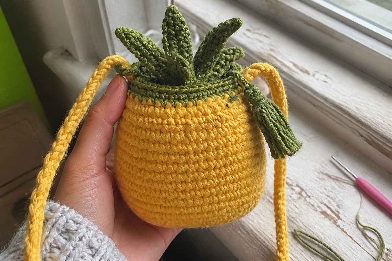 Yellow crocheted pineapple bag with green leaves held beside purple hook