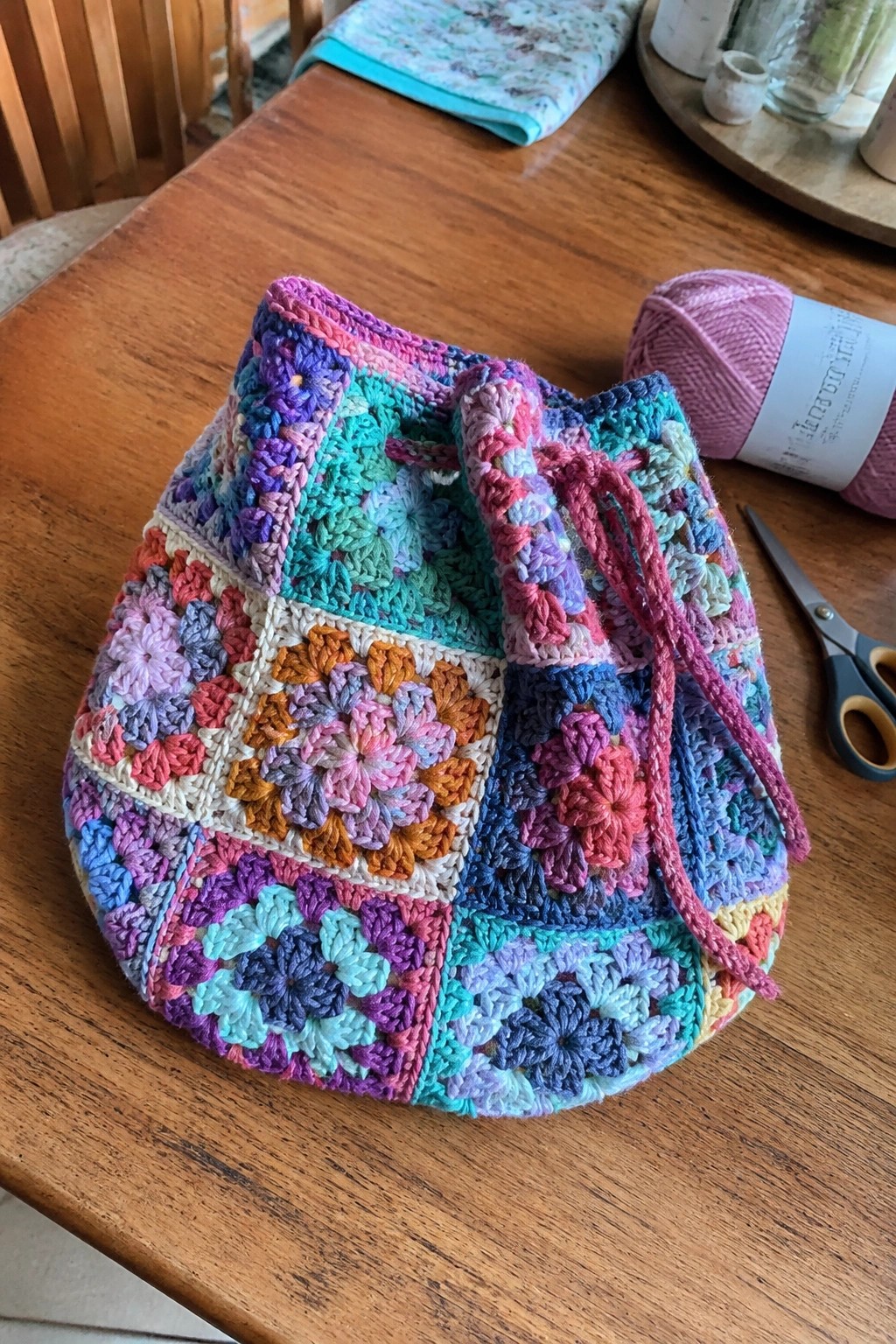 Colorful crocheted granny square drawstring bag on wooden table with yarn and scissors.