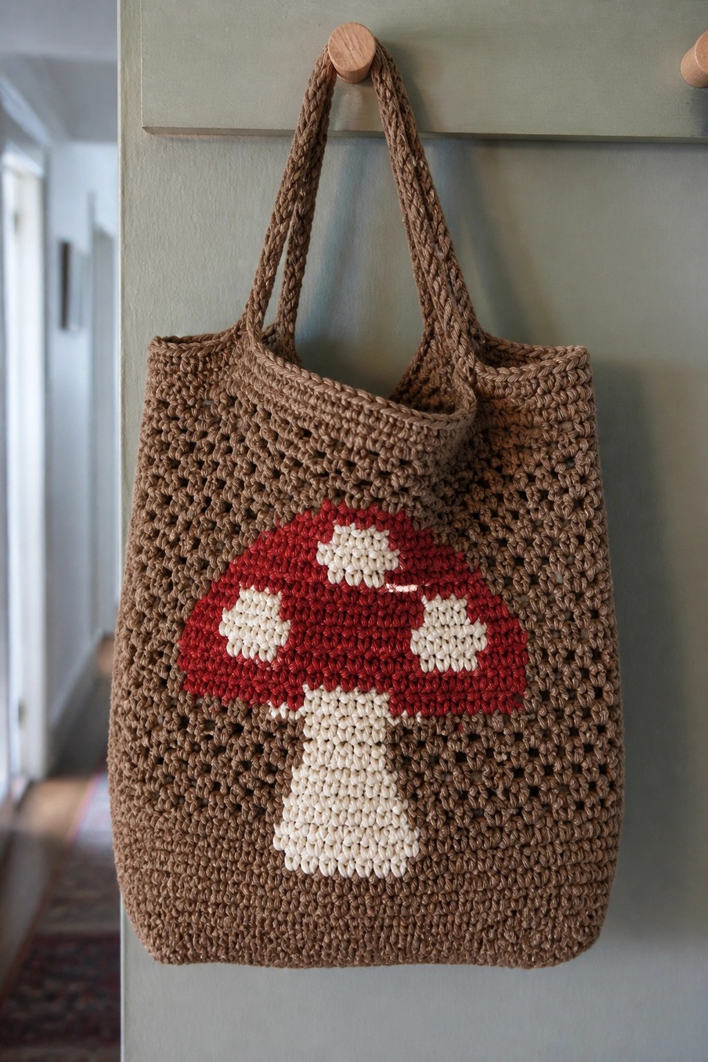 Brown crocheted tote bag with red mushroom design hanging on wooden hook.