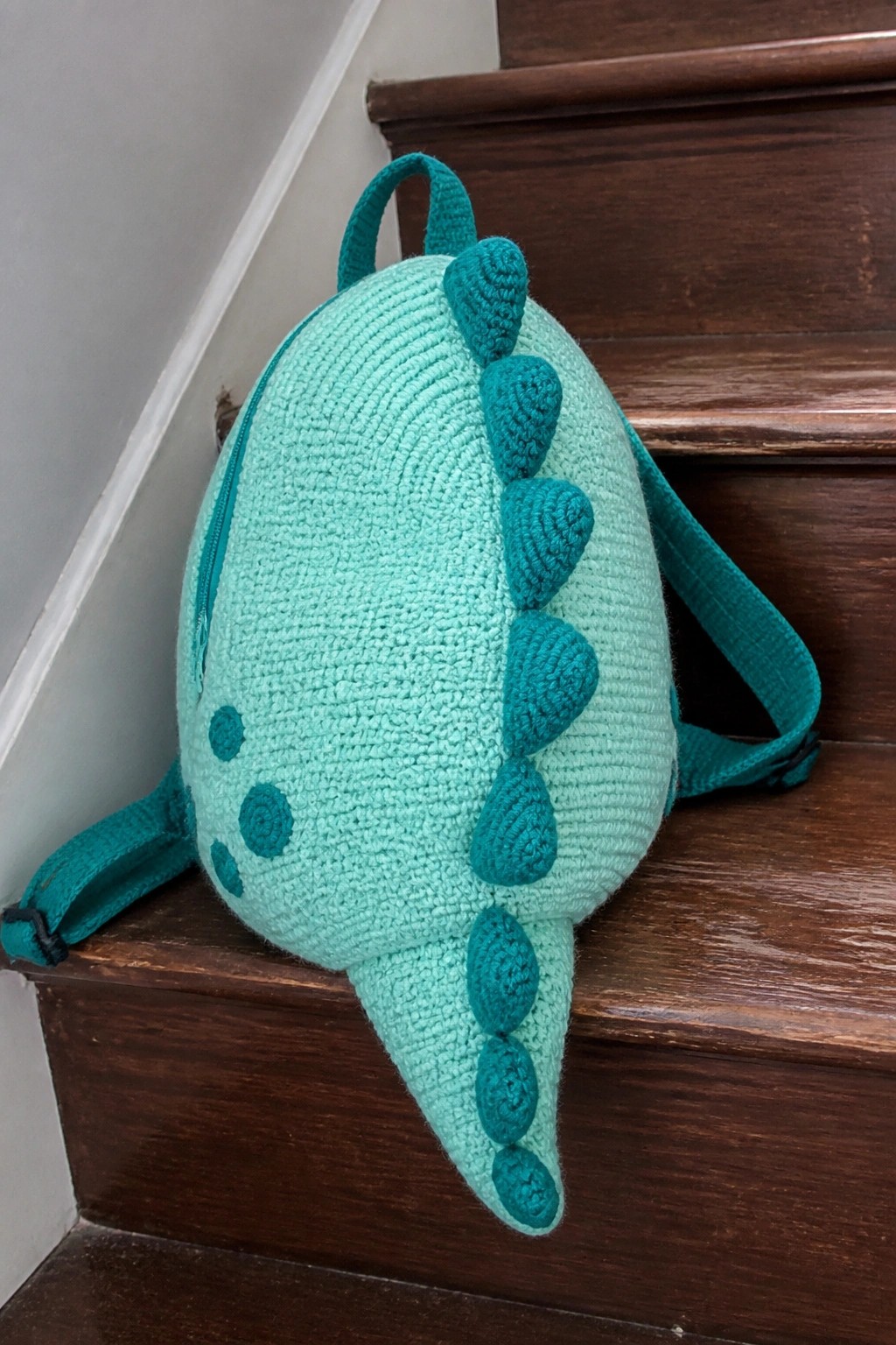 Mint green crocheted dinosaur backpack with teal spikes on wooden stairs.
