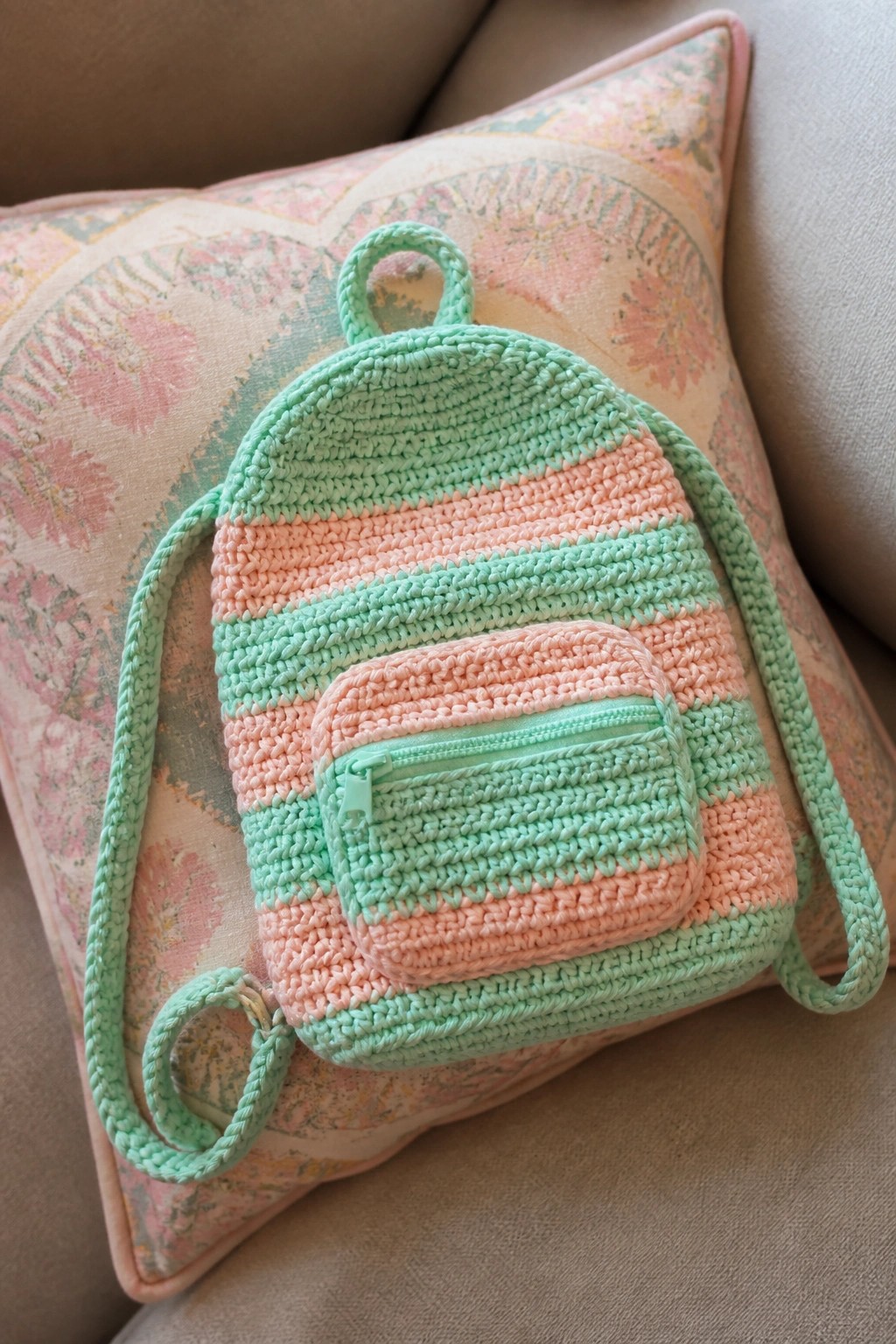 Mint green and peach striped crocheted backpack with zipper pocket on floral pillow.