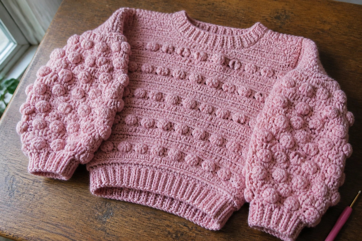 Pink bobble-stitch crochet sweater on wooden table beside hook.