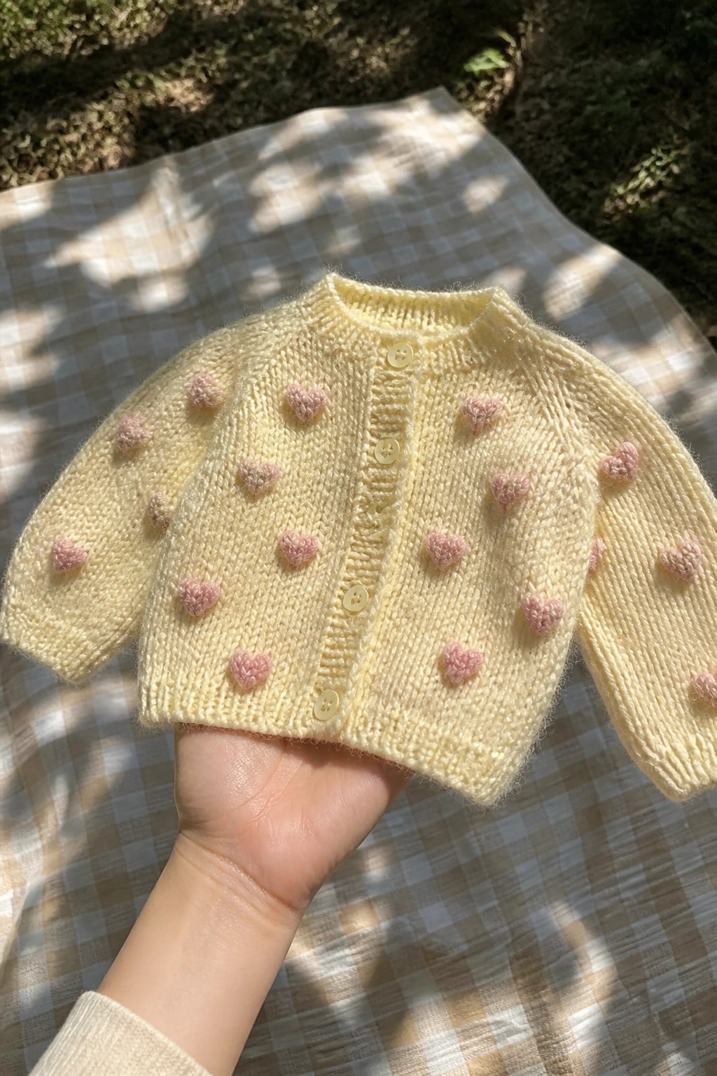 Yellow knitted baby cardigan with pink hearts held over checkered blanket