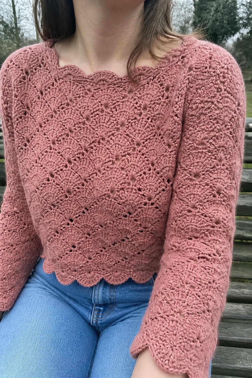 Pink crocheted lace sweater with scalloped edges over blue jeans on a bench