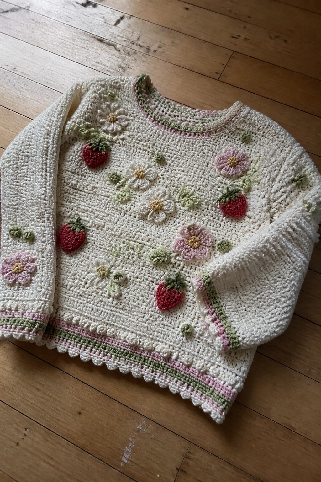 A cream crocheted sweater with strawberry and flower appliqués and striped borders lies flat on a wooden floor.