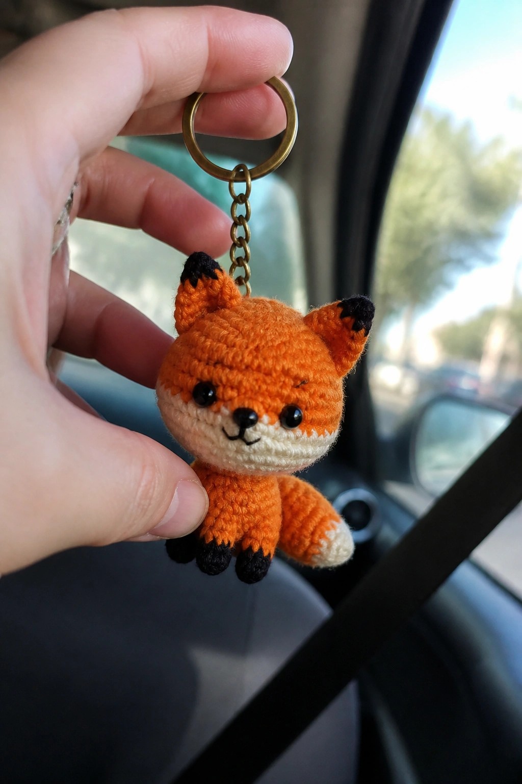 A hand holds a small crocheted orange fox keychain with black accents inside a car.