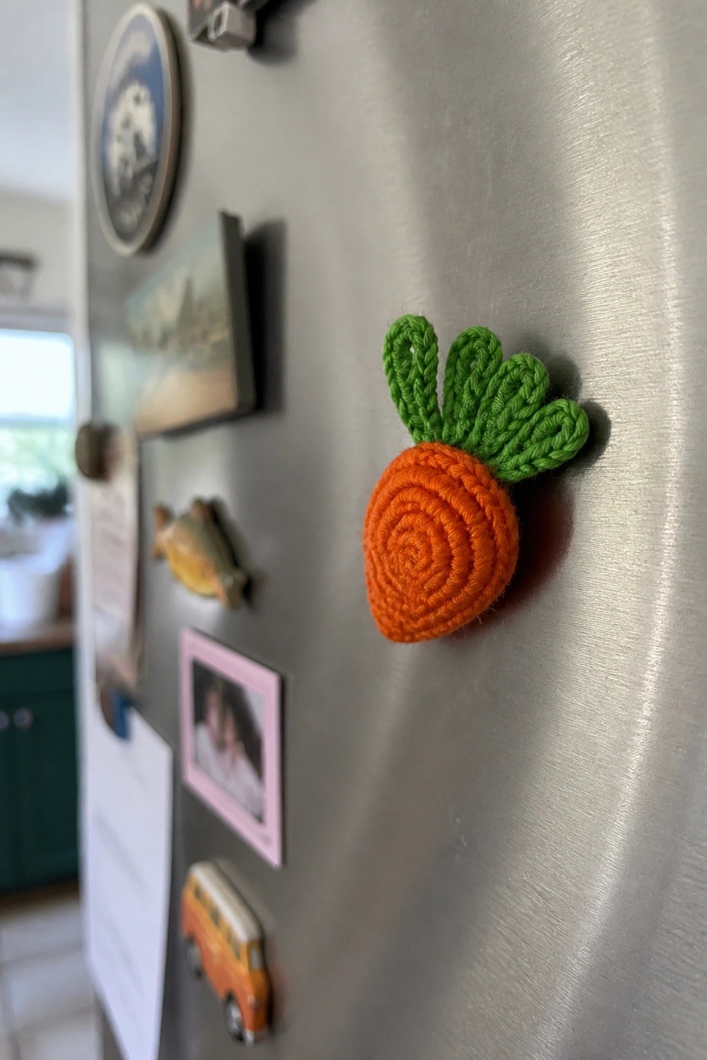 A crocheted orange carrot with a green top stuck to a stainless steel refrigerator door.