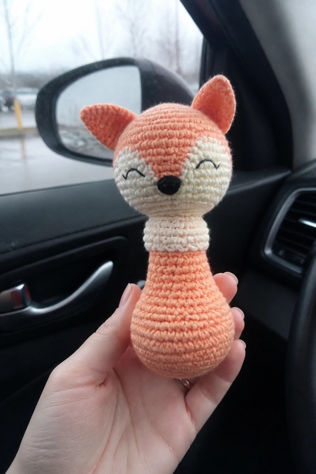 Hand holding crocheted peach fox with closed eyes inside a car.