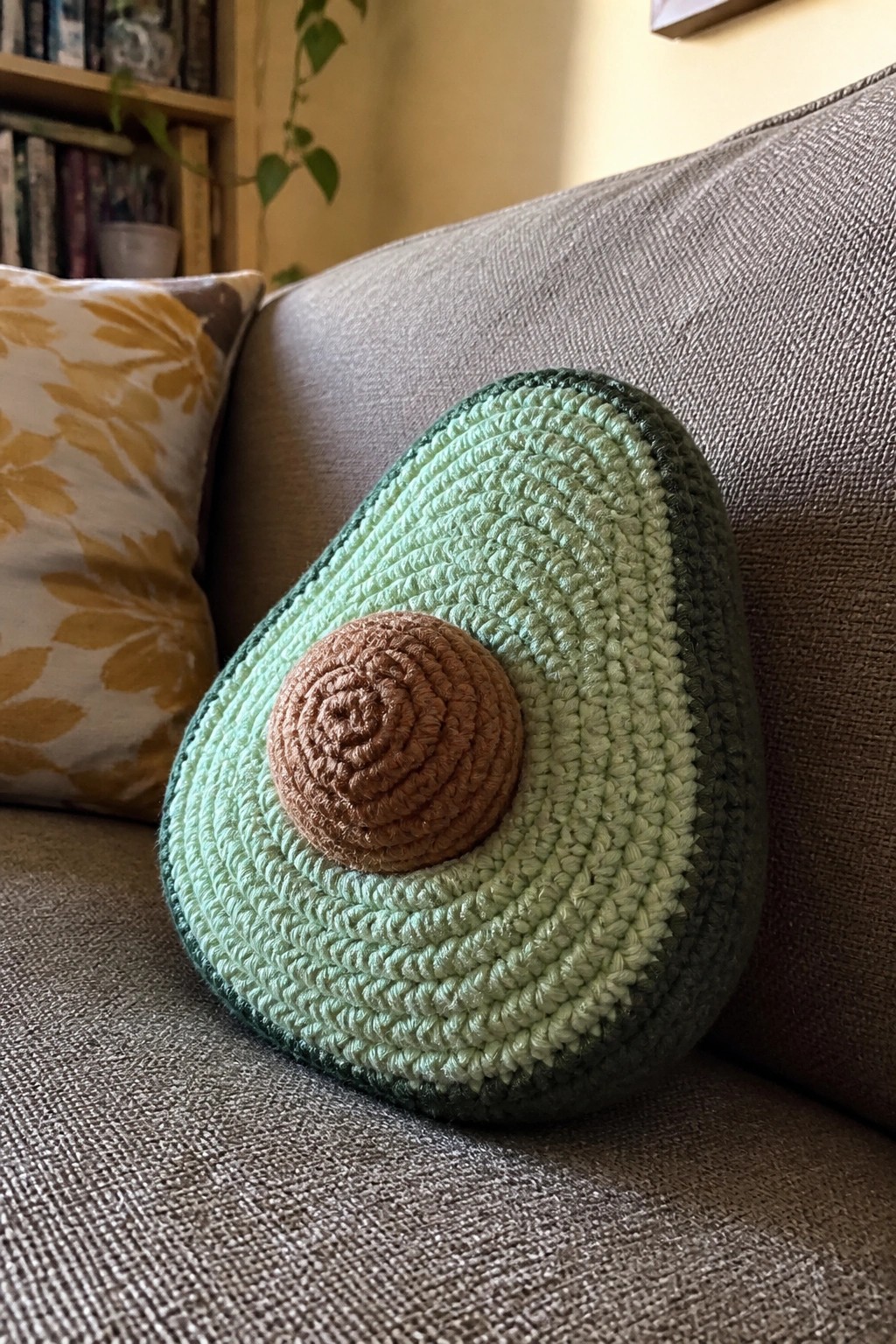 Crocheted avocado pillow on gray couch beside yellow patterned cushion