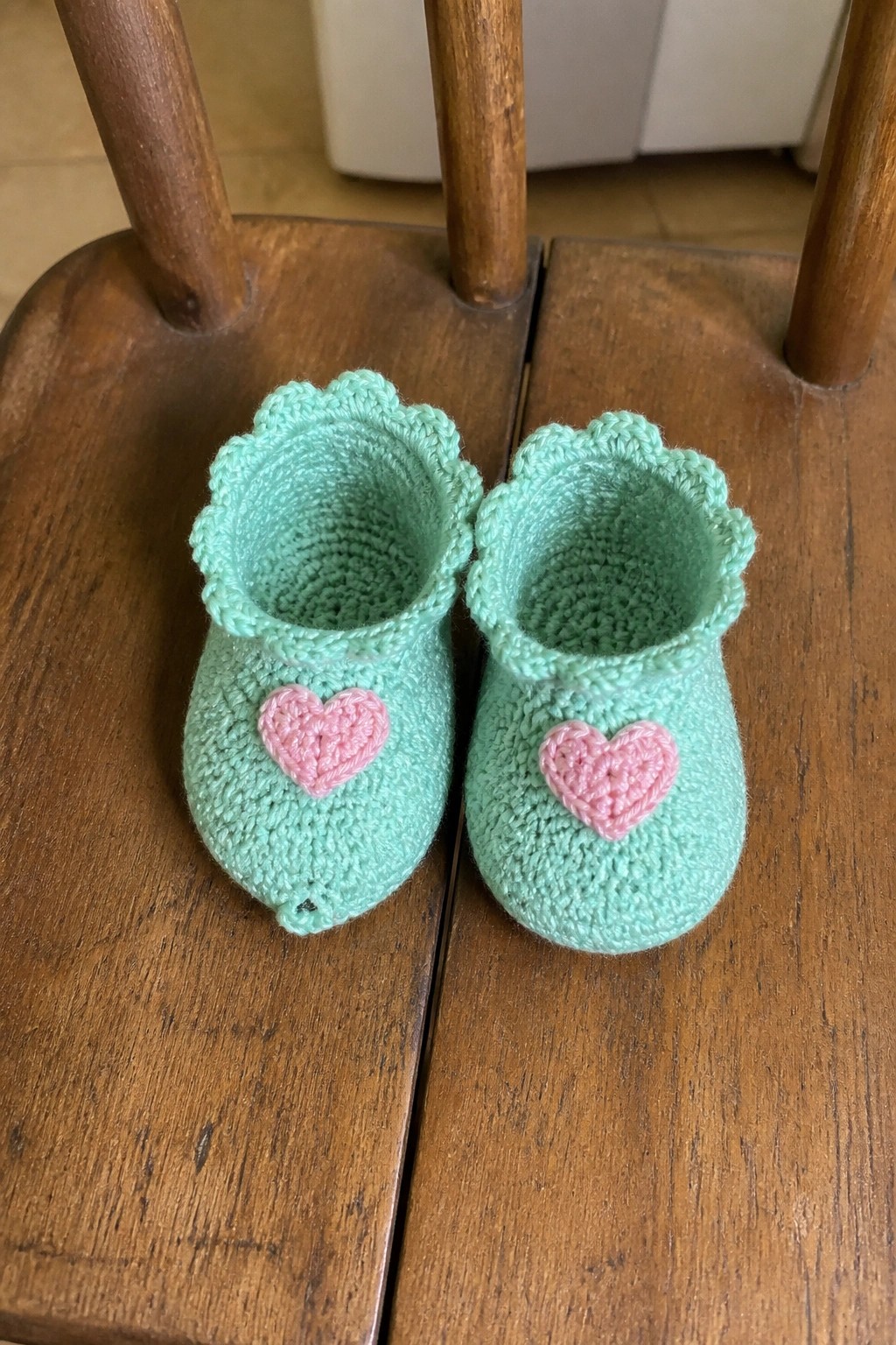 Mint green crocheted baby booties with pink hearts on wooden chair.