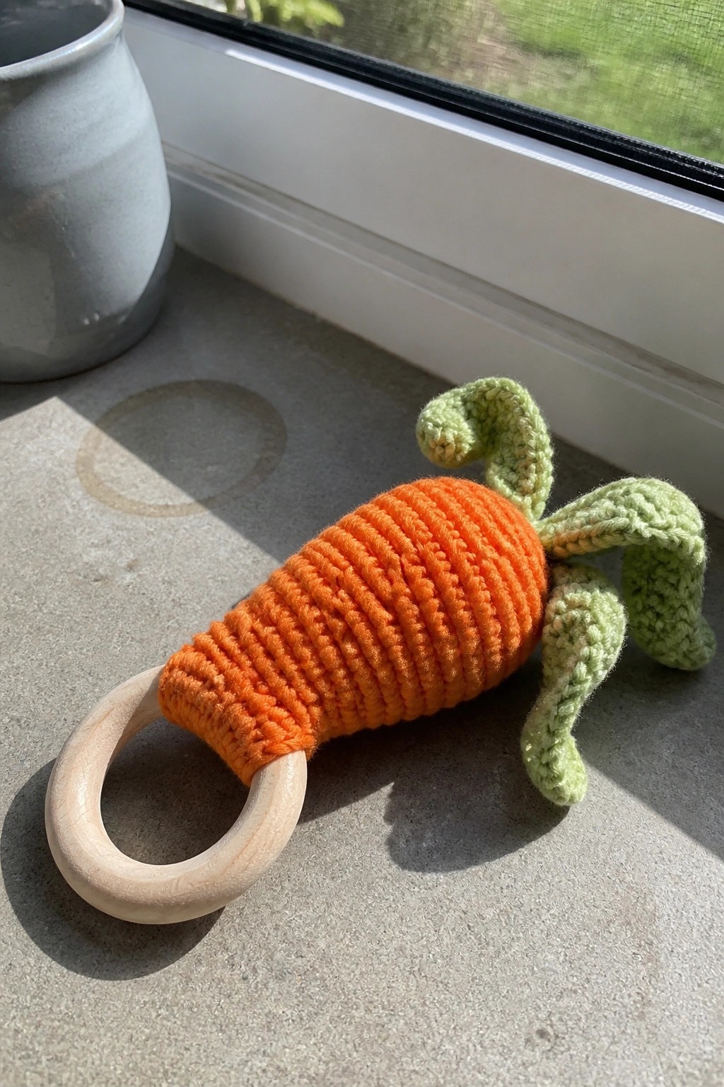 Orange crocheted carrot with green leaves attached to a wooden ring on a stone surface.