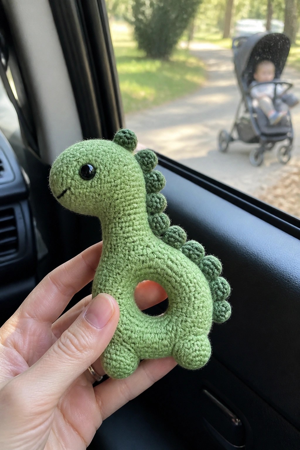 Hand holding green crocheted dinosaur rattle in car near stroller outside.