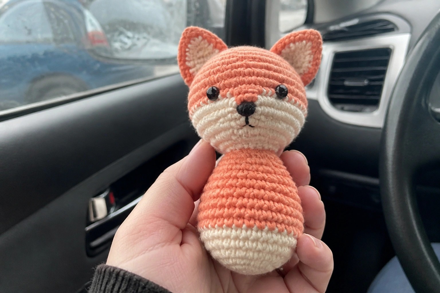 Hand holding crocheted peach fox with closed eyes inside a car.
