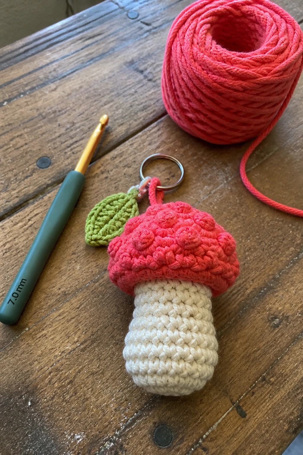 Crocheted pink mushroom keychain with white stem, hook, and yarn on wood table.