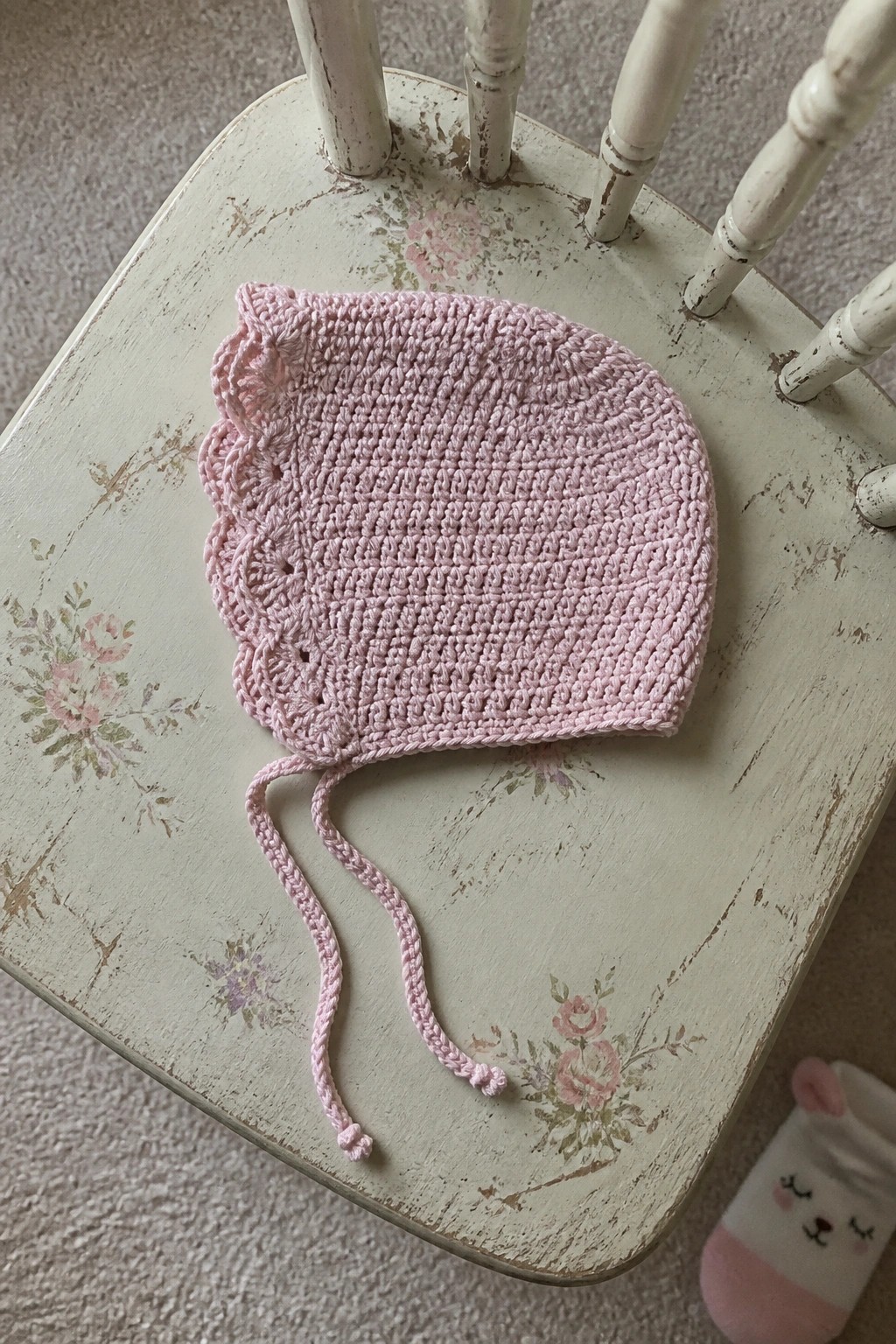 A pink crocheted baby bonnet with scalloped edges and ties rests on a white wooden chair.