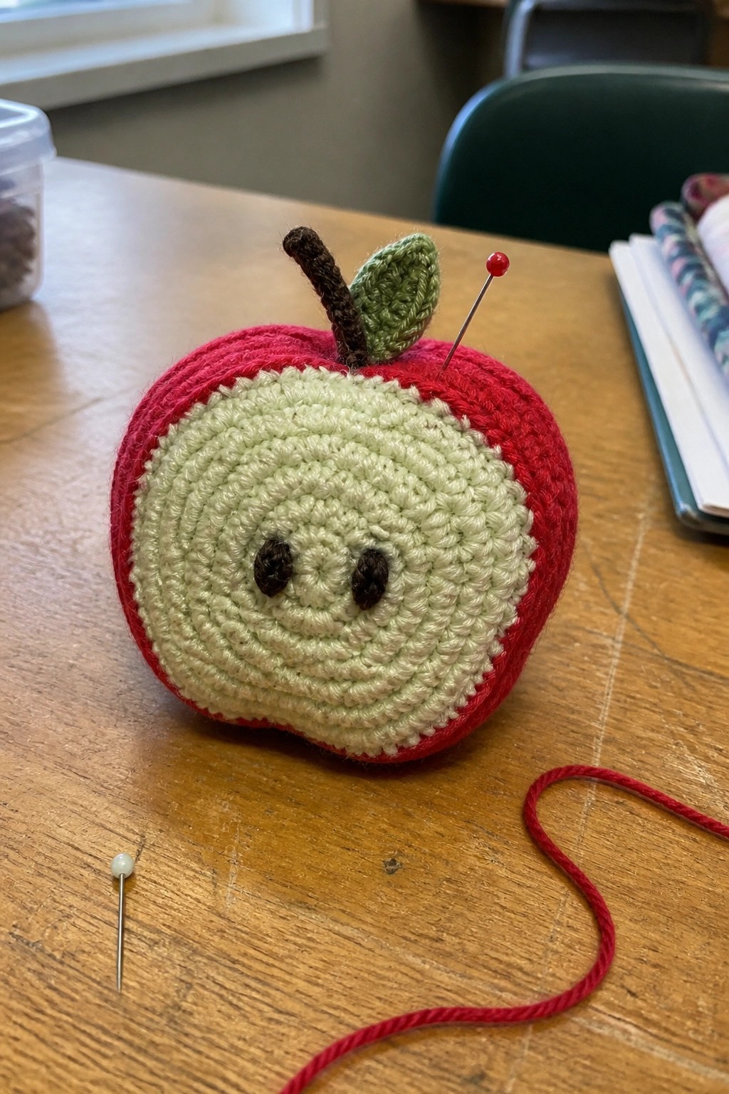 Crocheted red apple pincushion with green face, stem, and pins on table