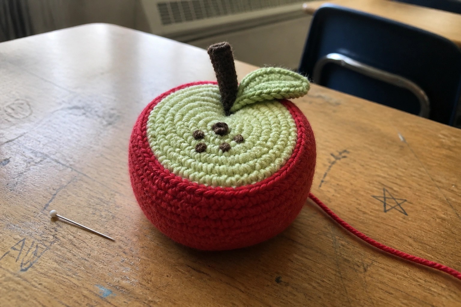 Crocheted red apple pincushion with green face, stem, and pins on table