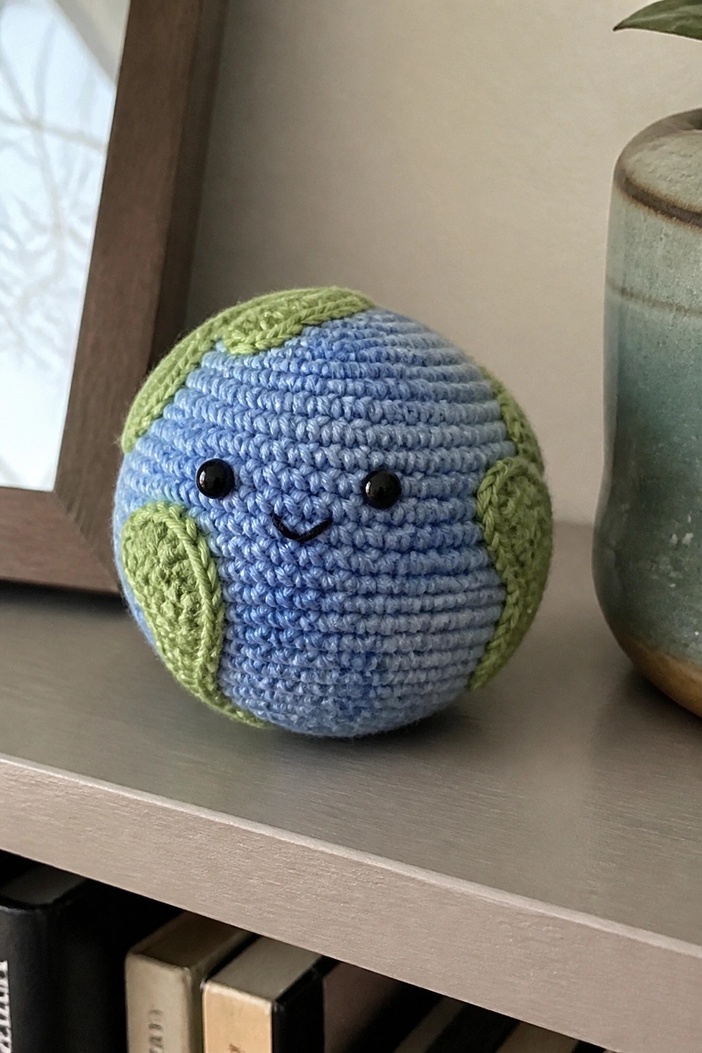 Crocheted blue Earth plush with green patches and smiling face on wooden shelf