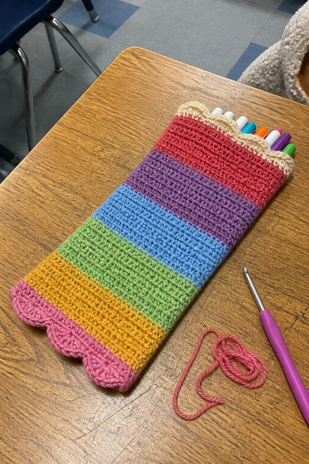 A rainbow striped crocheted pouch holding colorful markers on a wooden desk next to a crochet hook and pink yarn.
