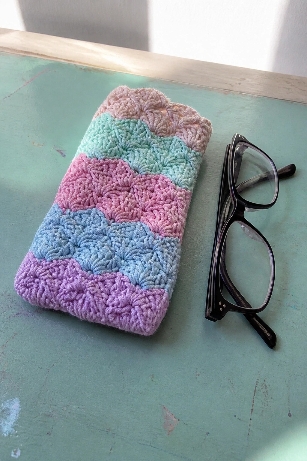 A crocheted rectangular pouch with horizontal pastel stripes in pink, mint, blue, and purple rests on a green surface next to a pair of black glasses.
