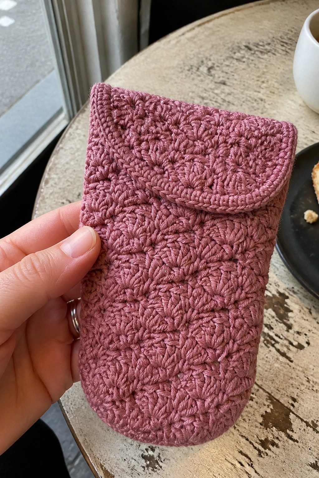 Hand holding a dusty pink crocheted pouch with flap on wooden table