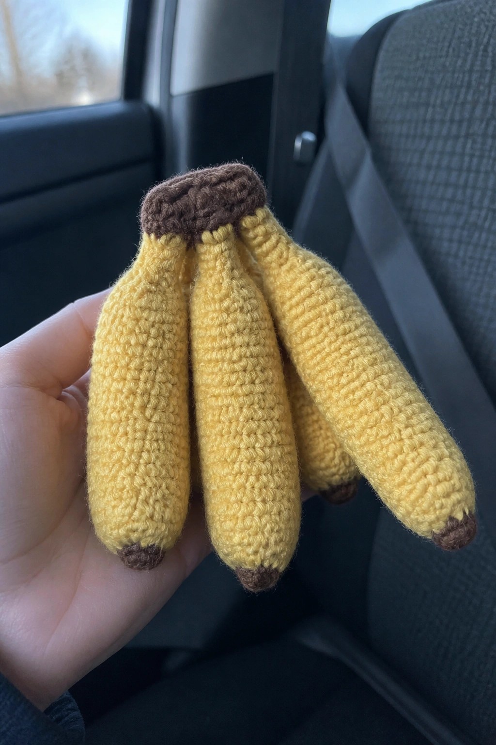 A hand holds a small crocheted bunch of yellow bananas with brown tips inside a car.