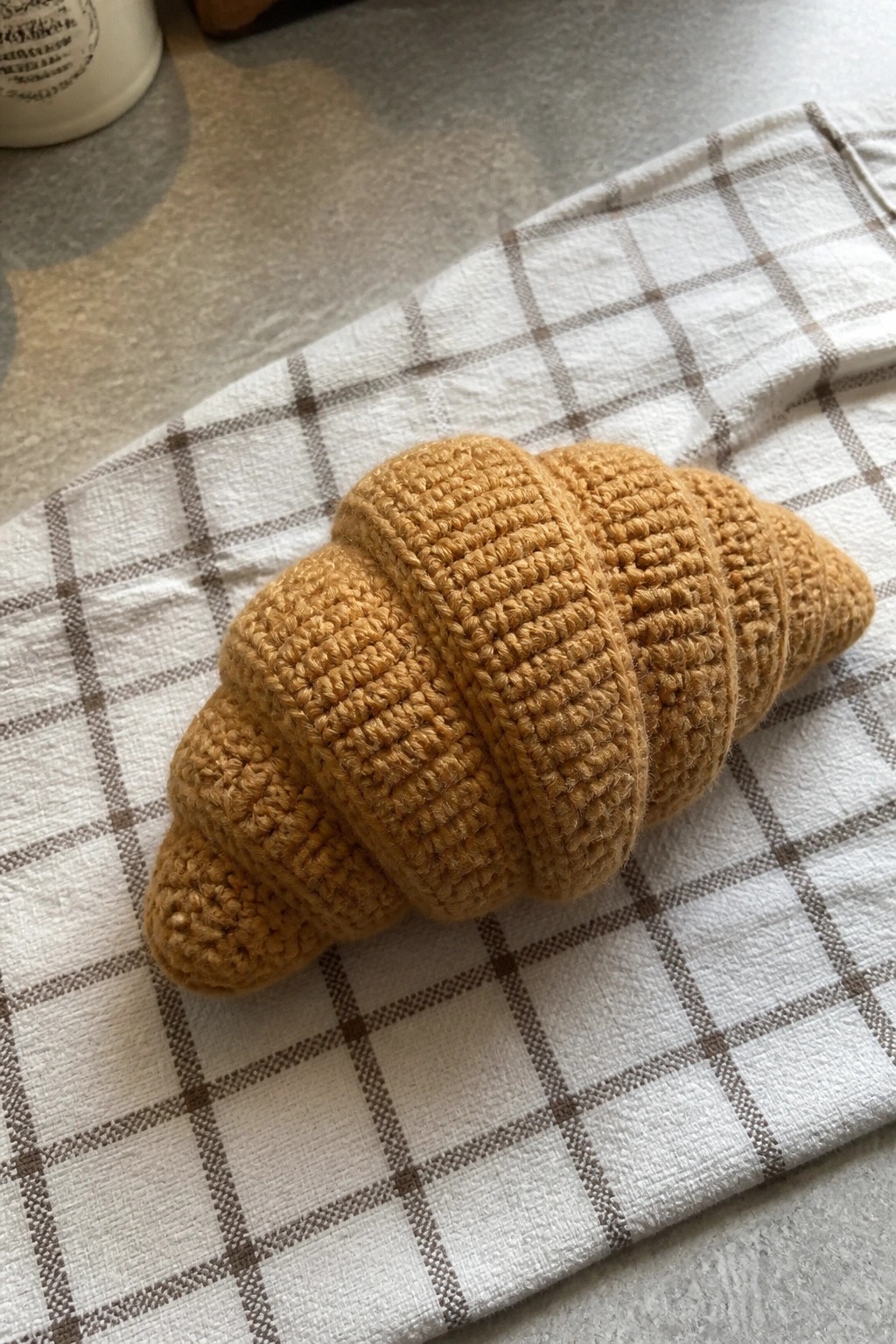 A golden-brown crocheted croissant plushie on a white and brown checkered cloth.