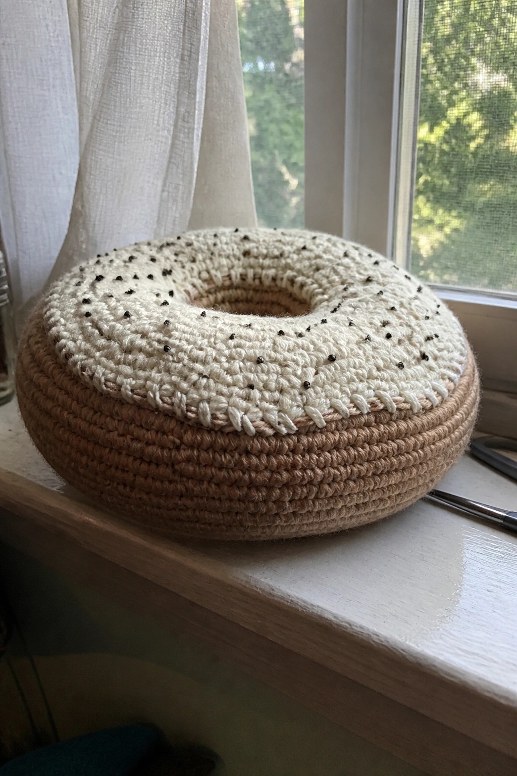Crocheted donut pillow in brown and cream with black dots on windowsill