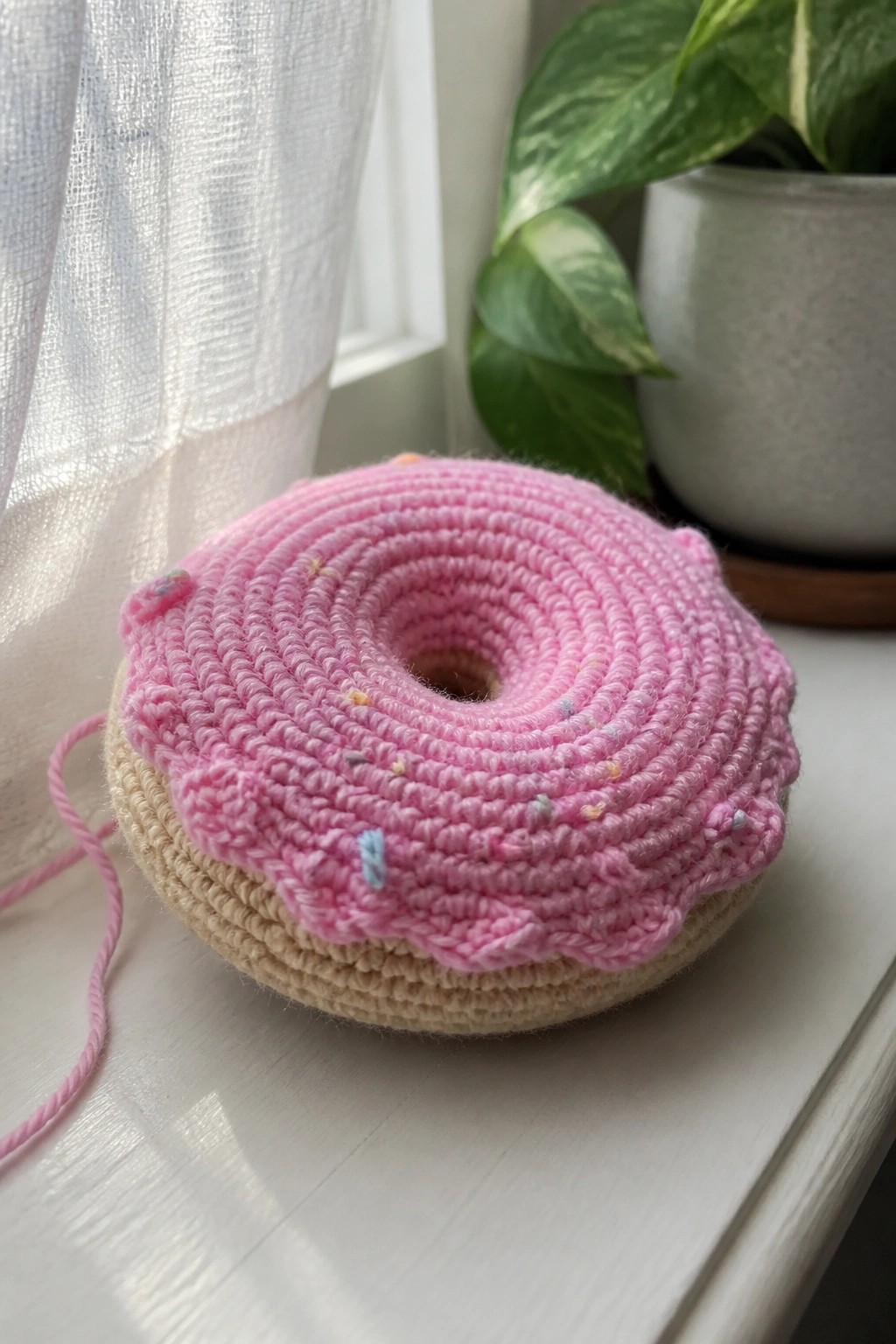 Pink crocheted donut with sprinkles on beige base atop white windowsill near plant.