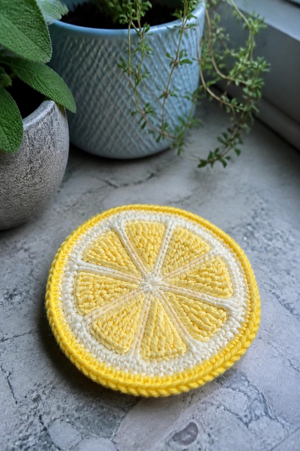 A round crocheted coaster in yellow and white lemon slice design rests on a gray countertop near potted plants.