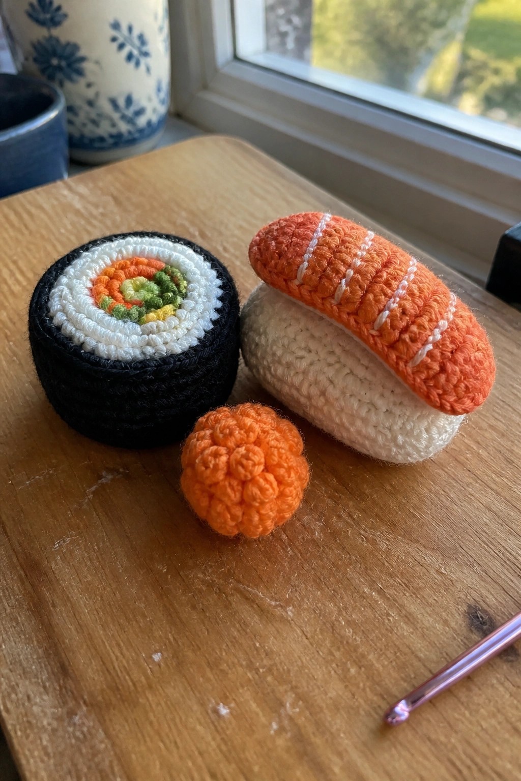 Crocheted maki roll, nigiri, and roe with pink hook on wooden table by window
