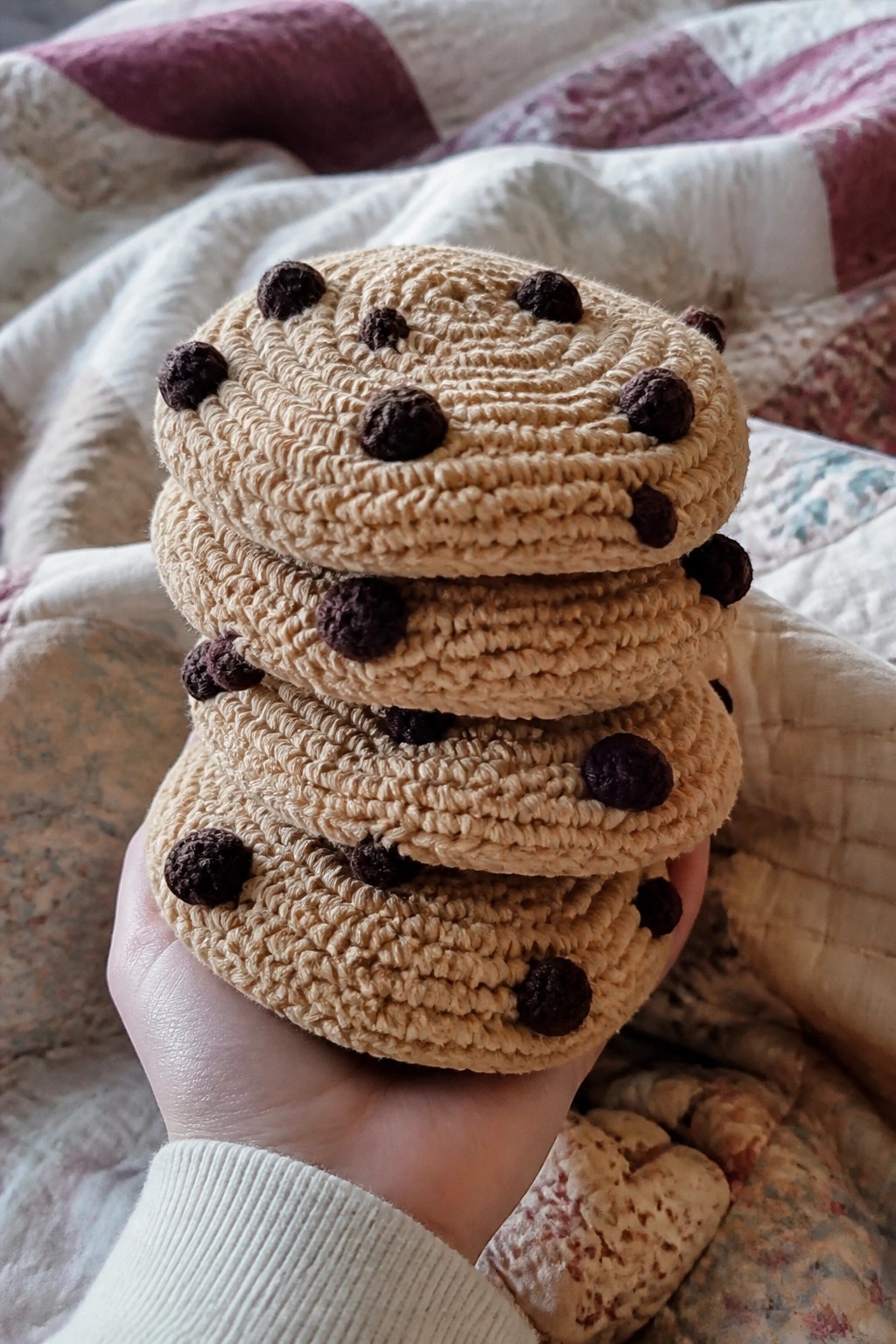 Hand holding stack of beige crocheted cookies with brown dots on quilt.