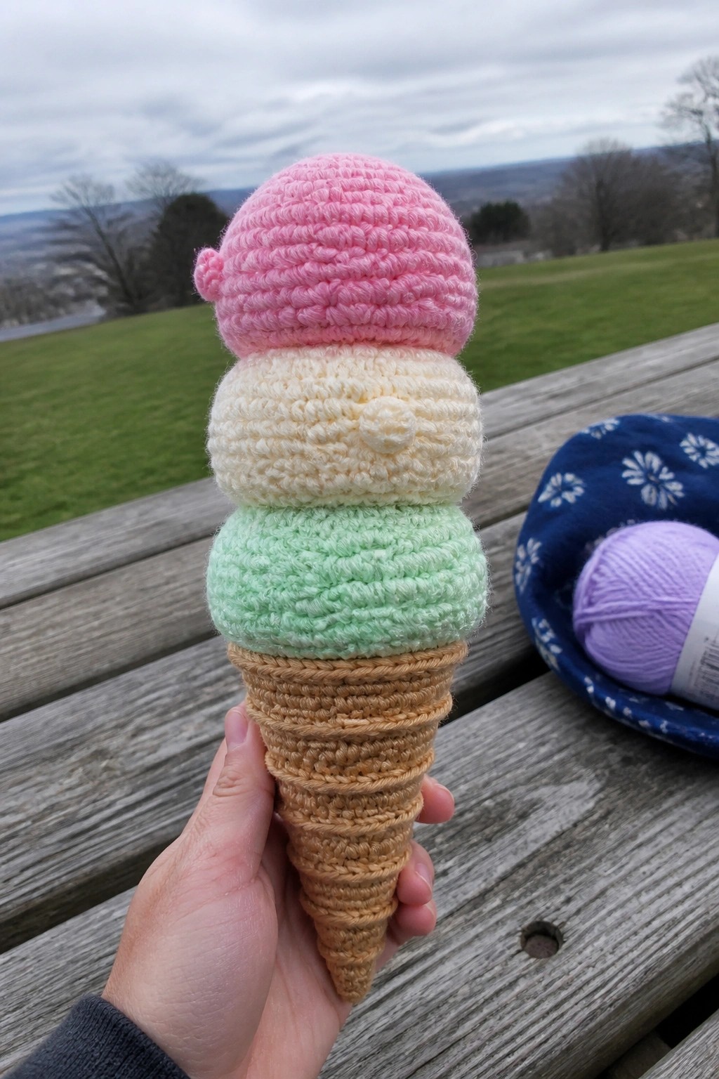 Hand holding crocheted ice cream cone with pink, cream, and mint scoops on bench