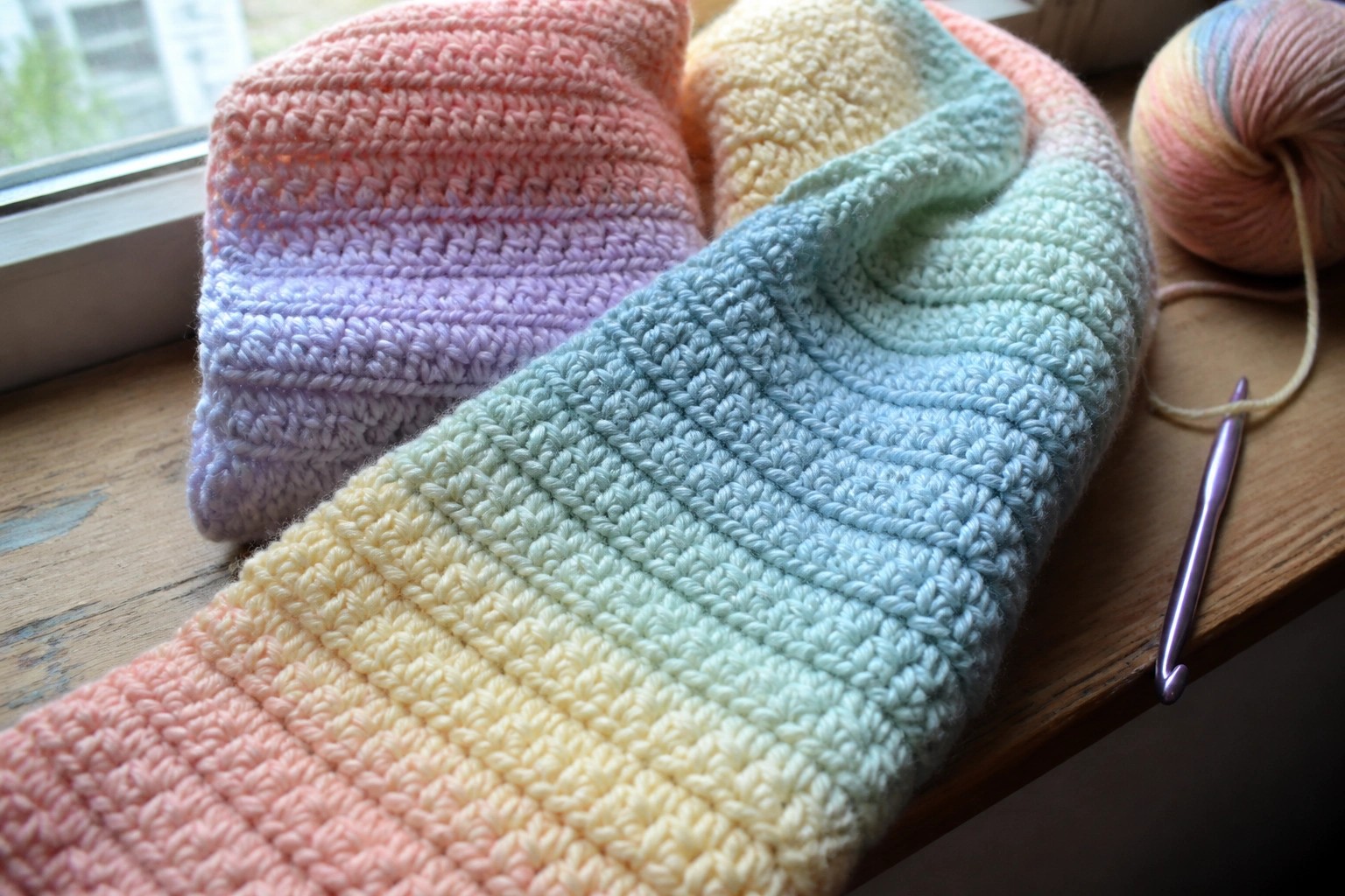 Pastel rainbow crocheted blanket on wooden windowsill with yarn and hook