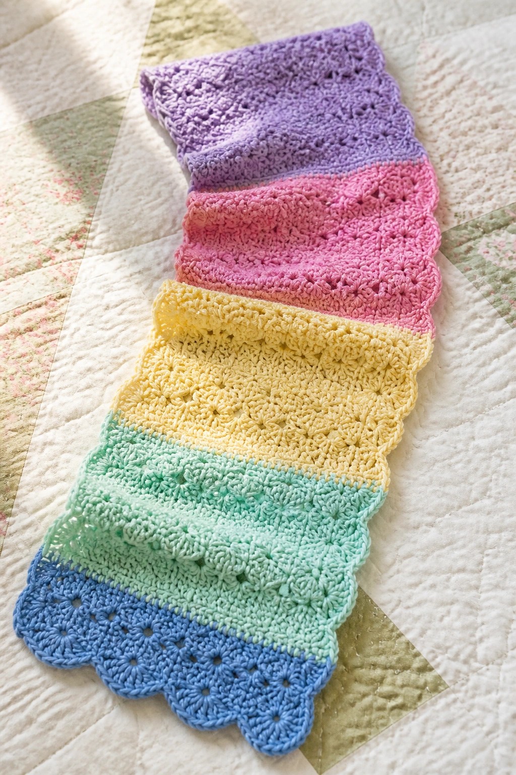 Pastel rainbow crocheted scarf with scalloped edges on floral quilt.