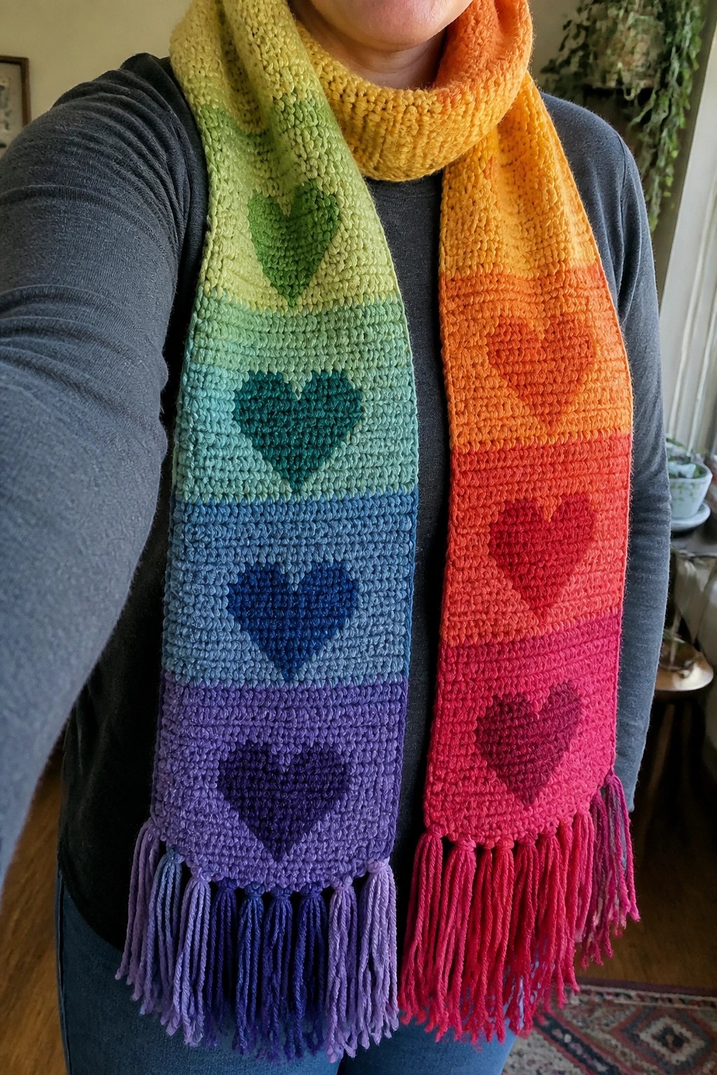 A person wearing a long crocheted scarf with rainbow gradient stripes and one heart motif per stripe, finished with color-matched fringes.